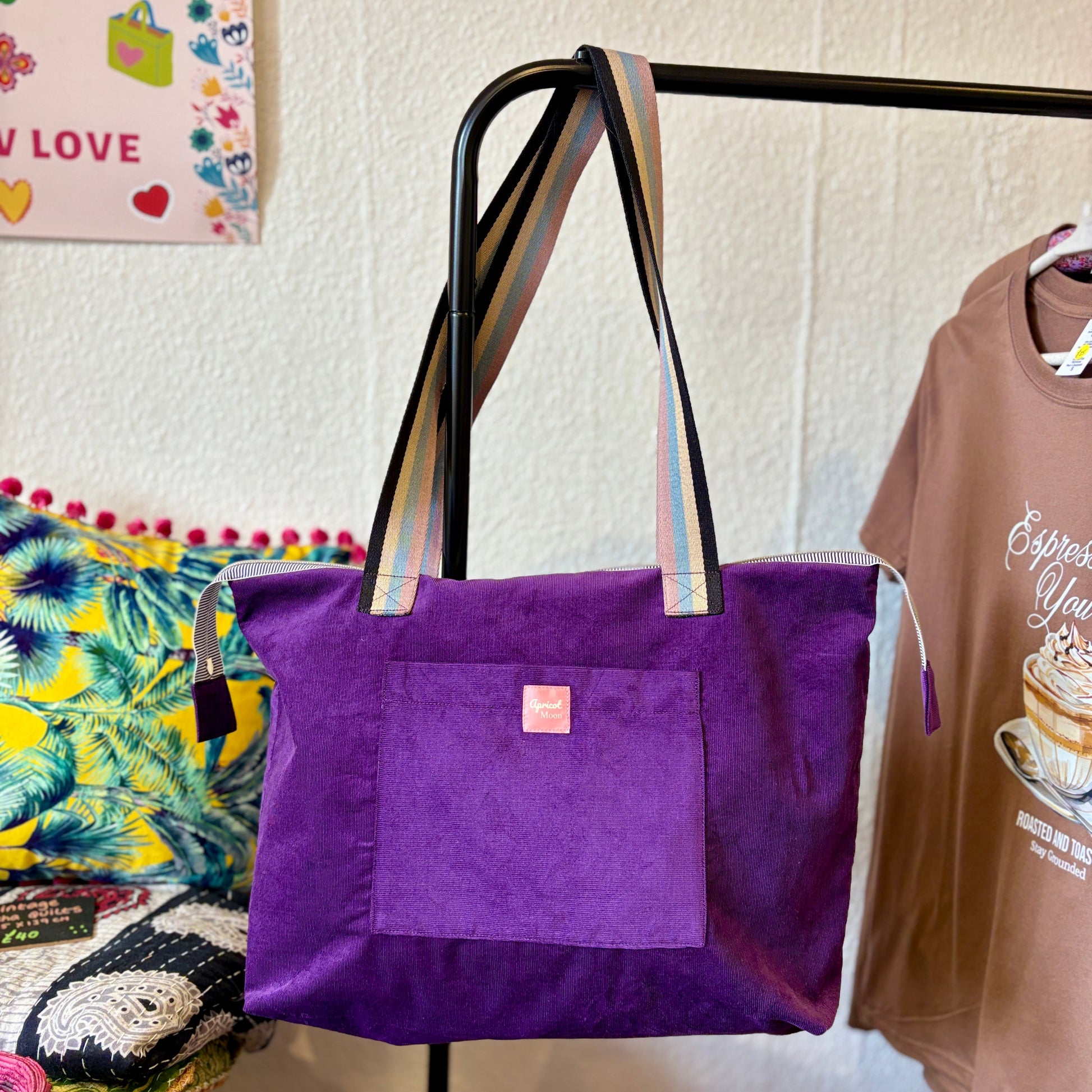 Purple tote bag with striped handles hanging on a rack next to a brown shirt.