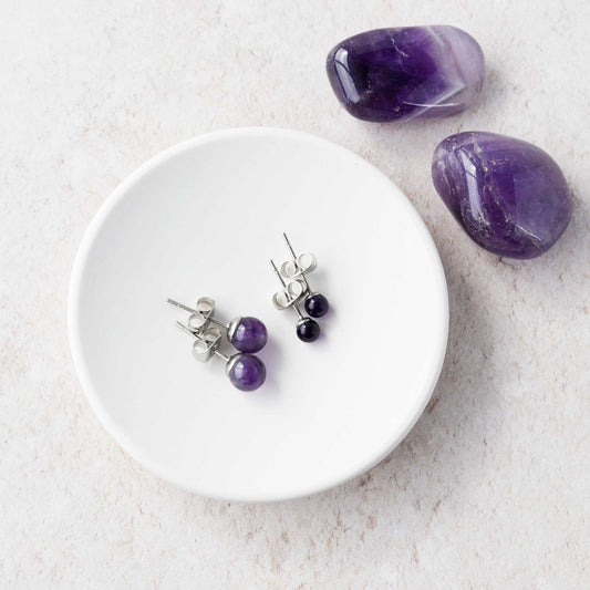 Pair of purple beaded earrings on a white plate with two purple stones on a light gray background