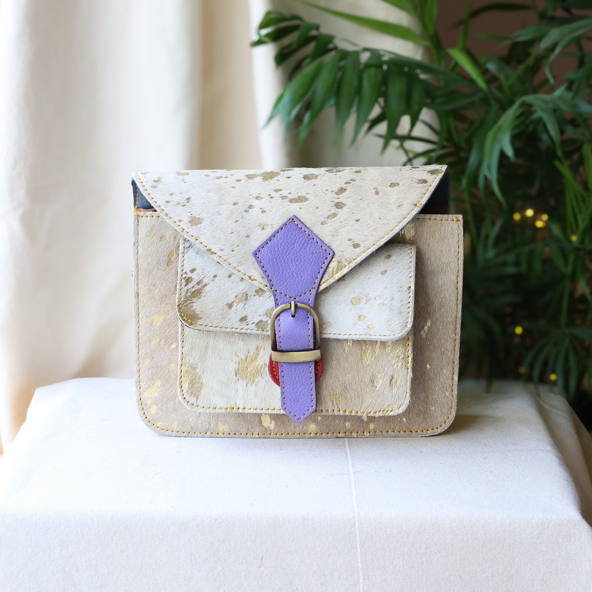 Small beige handbag with a purple strap on a white surface with a plant in the background