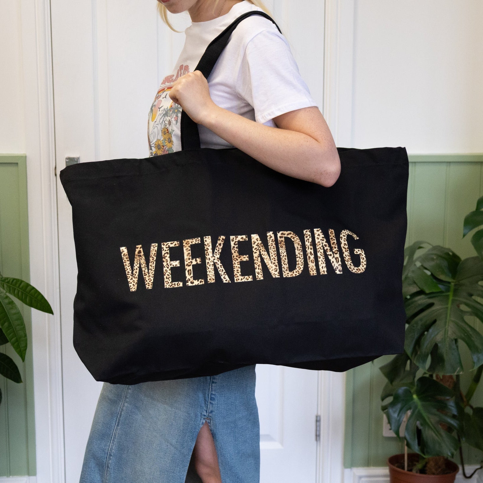 Person holding a black tote bag with 'WEEKENDING' text, standing indoors with plants in the background.