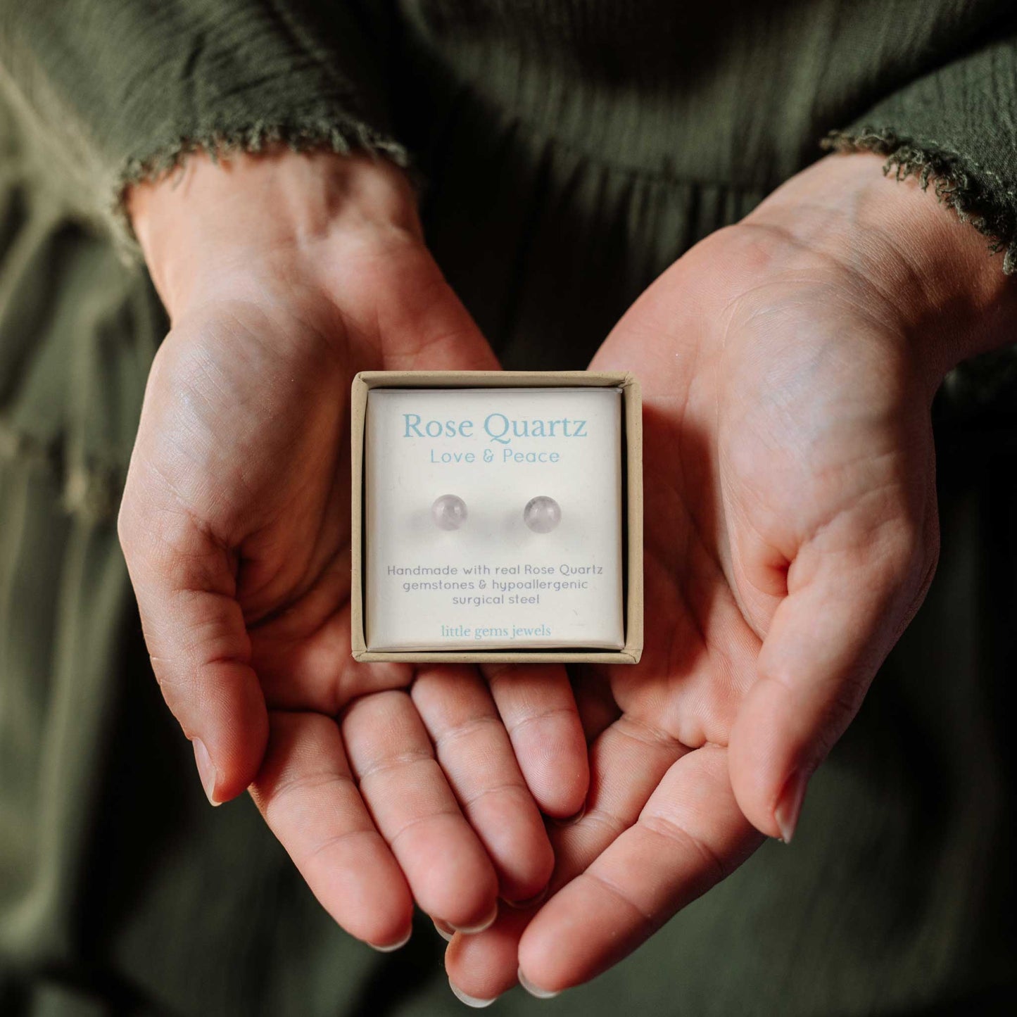 Hands holding small square eco friendly gift box containing Rose Quartz gemstone ball stud earrings.
