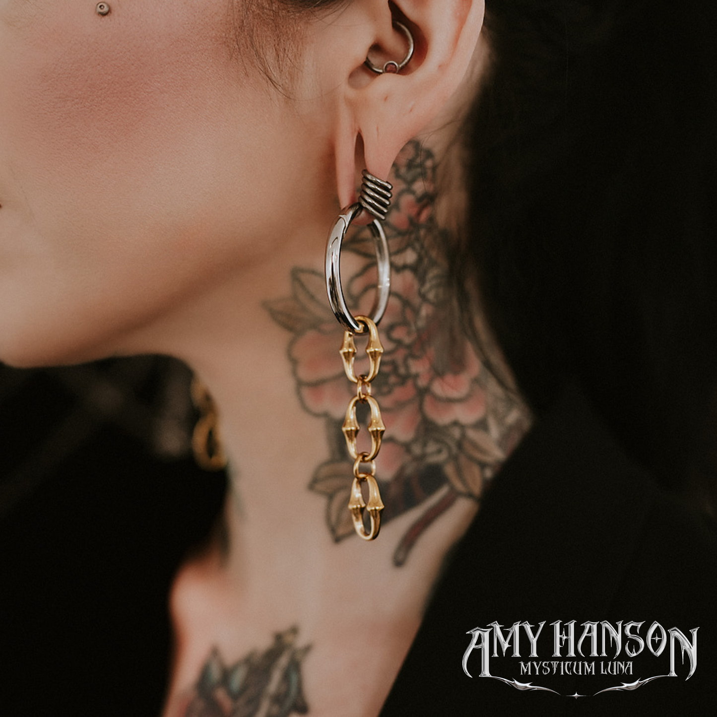 Close-up of an ear with a large hoop earring and tattoos on the neck, with 'Amy Hanson Mysticum Lumi' branding.
