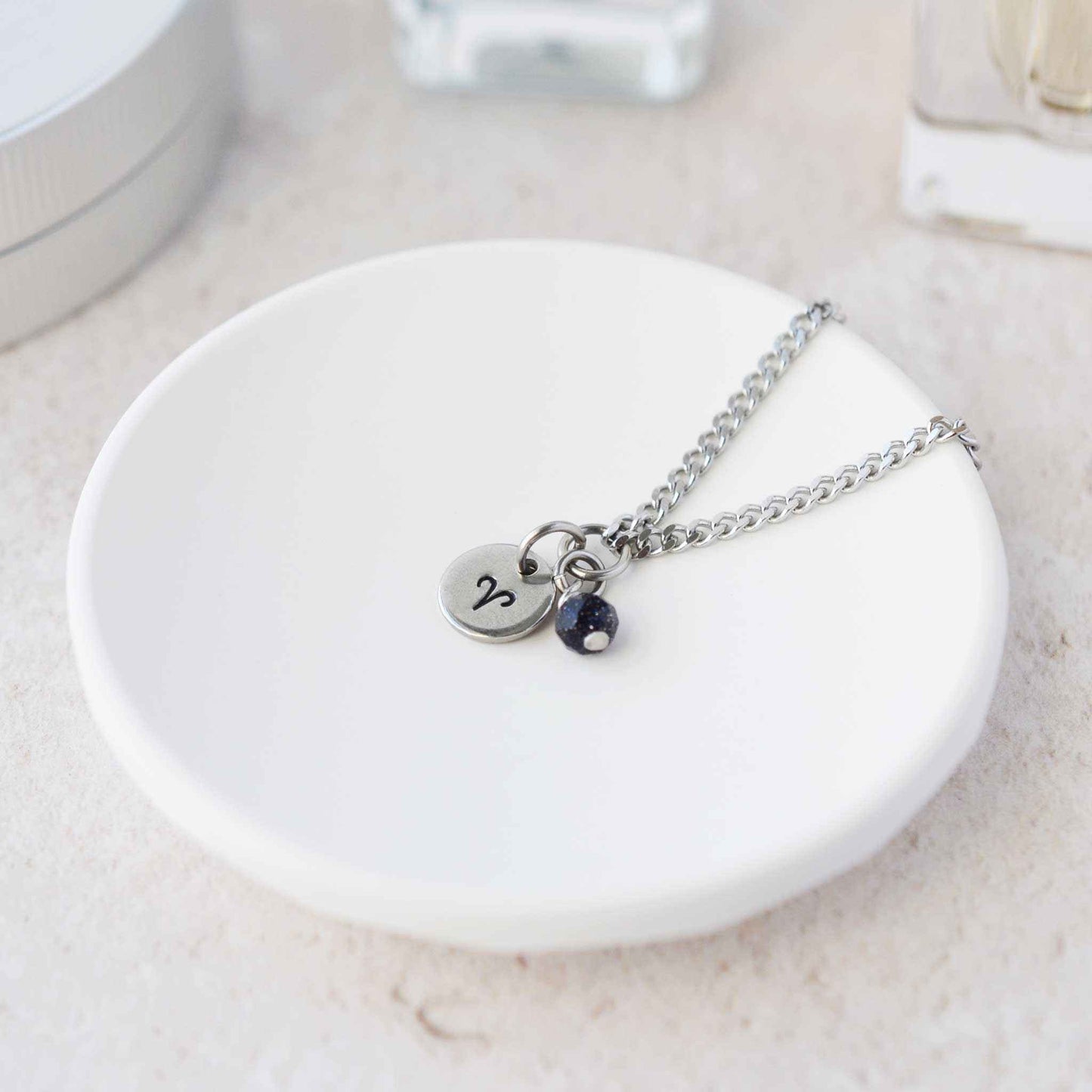 Tiny Aries zodiac sign disc pendant necklace with Blue Goldstone charm laying on white trinket dish