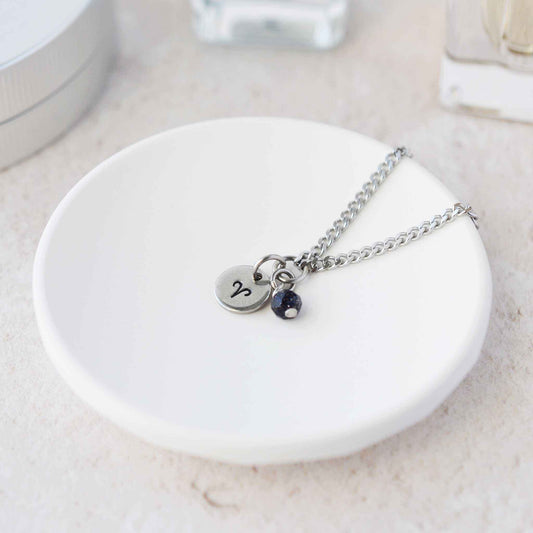 Tiny Aries zodiac sign disc pendant necklace with Blue Goldstone charm laying on white trinket dish