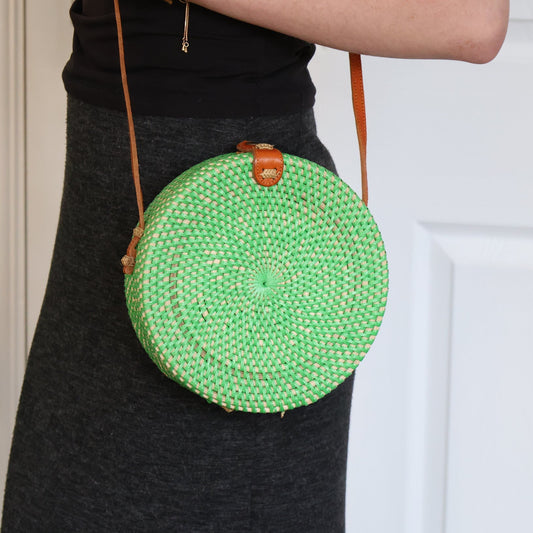 Green woven round bag with brown strap held by a person against a white door.