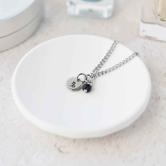 Tiny Capricorn zodiac sign disc pendant necklace with Blue Goldstone charm laying on white trinket dish