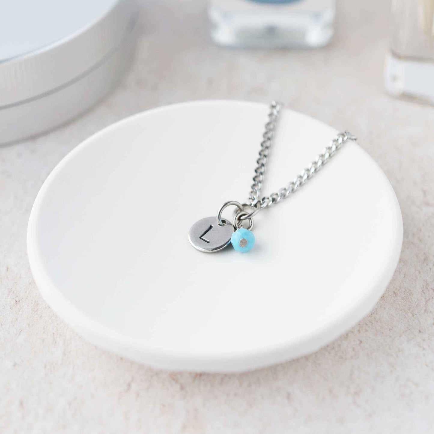 Tiny necklace with round disc pendant stamped with letter L and Turquoise gemstone charm laying in a white trinket dish