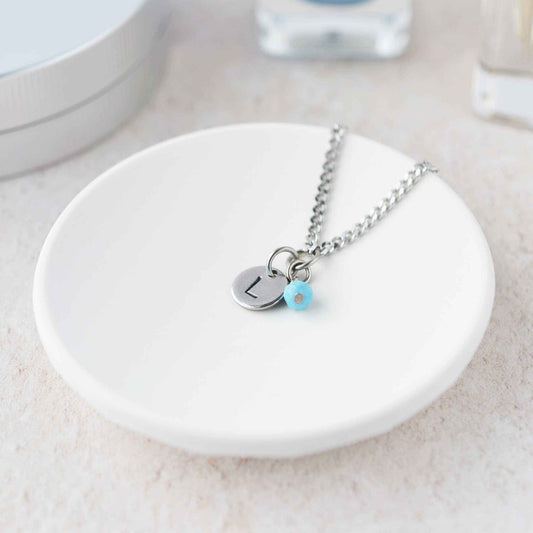 Tiny necklace with round disc pendant stamped with letter L and Turquoise gemstone charm laying in a white trinket dish