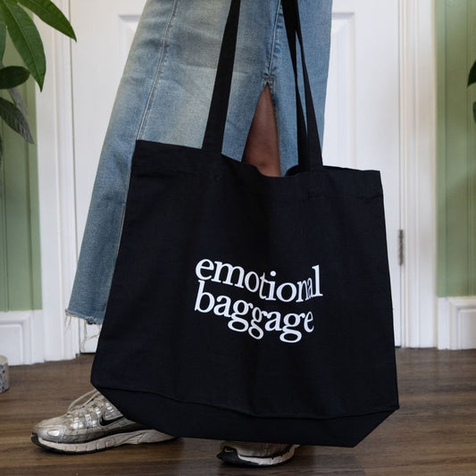 Person holding a black tote bag with 'emotional baggage' text, wearing a denim skirt indoors.