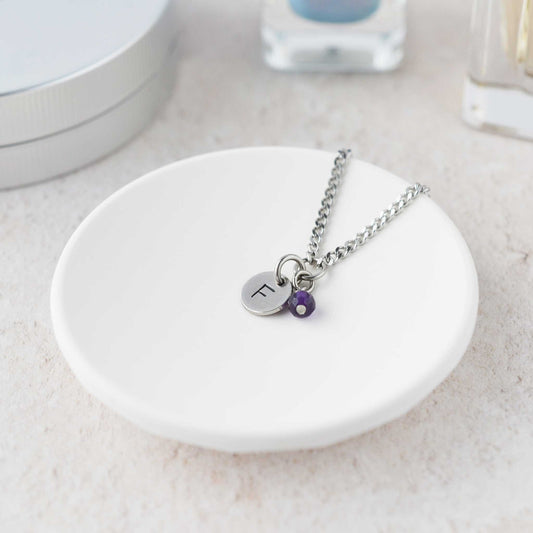 Tiny necklace with round disc pendant stamped with letter F and Amethyst gemstone charm laying in a white trinket dish