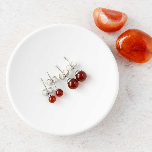 Two pairs of Carnelian stud earrings on a white trinket dish, with two red stones on a light surface.