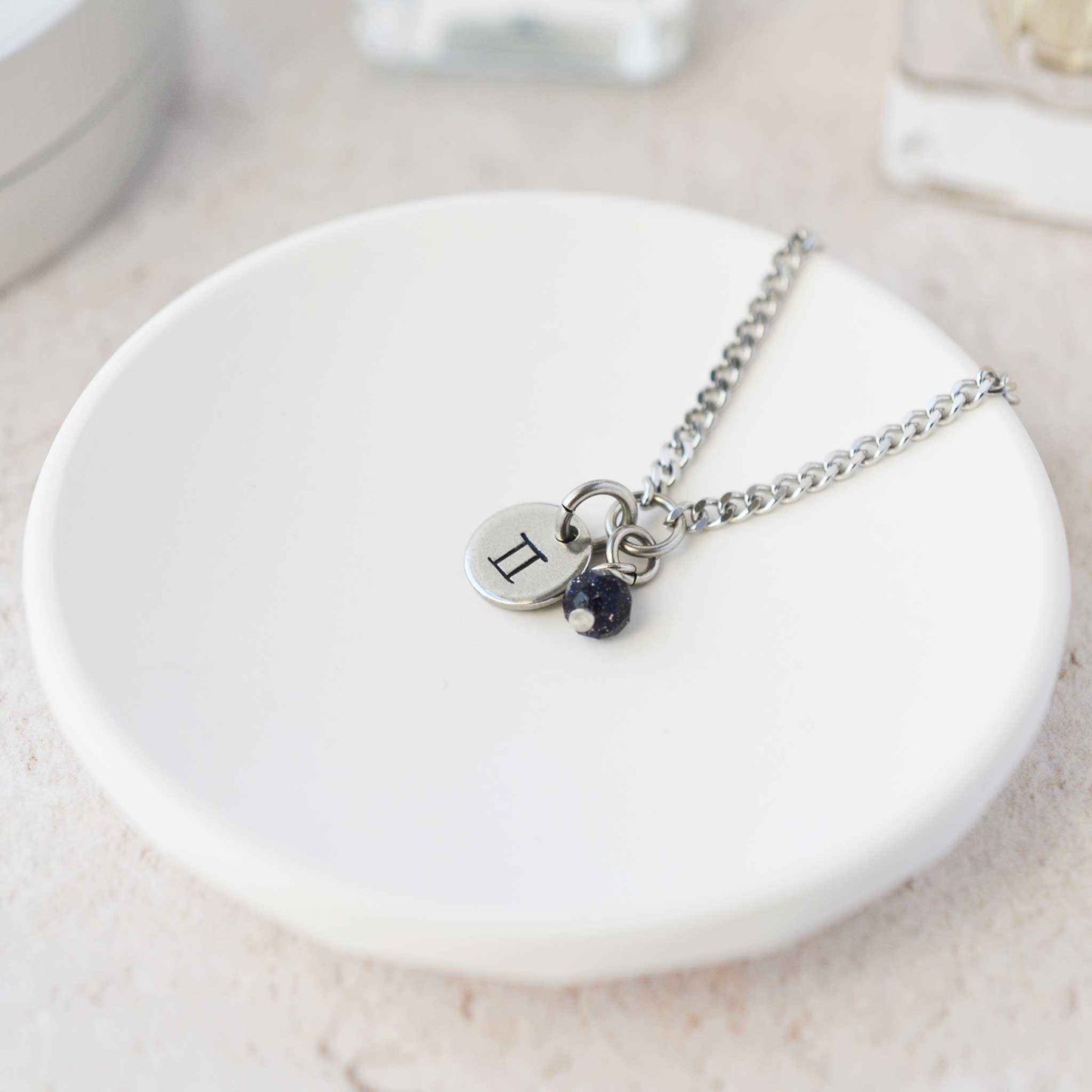 Tiny Gemini zodiac sign disc pendant necklace with Blue Goldstone charm laying on white trinket dish
