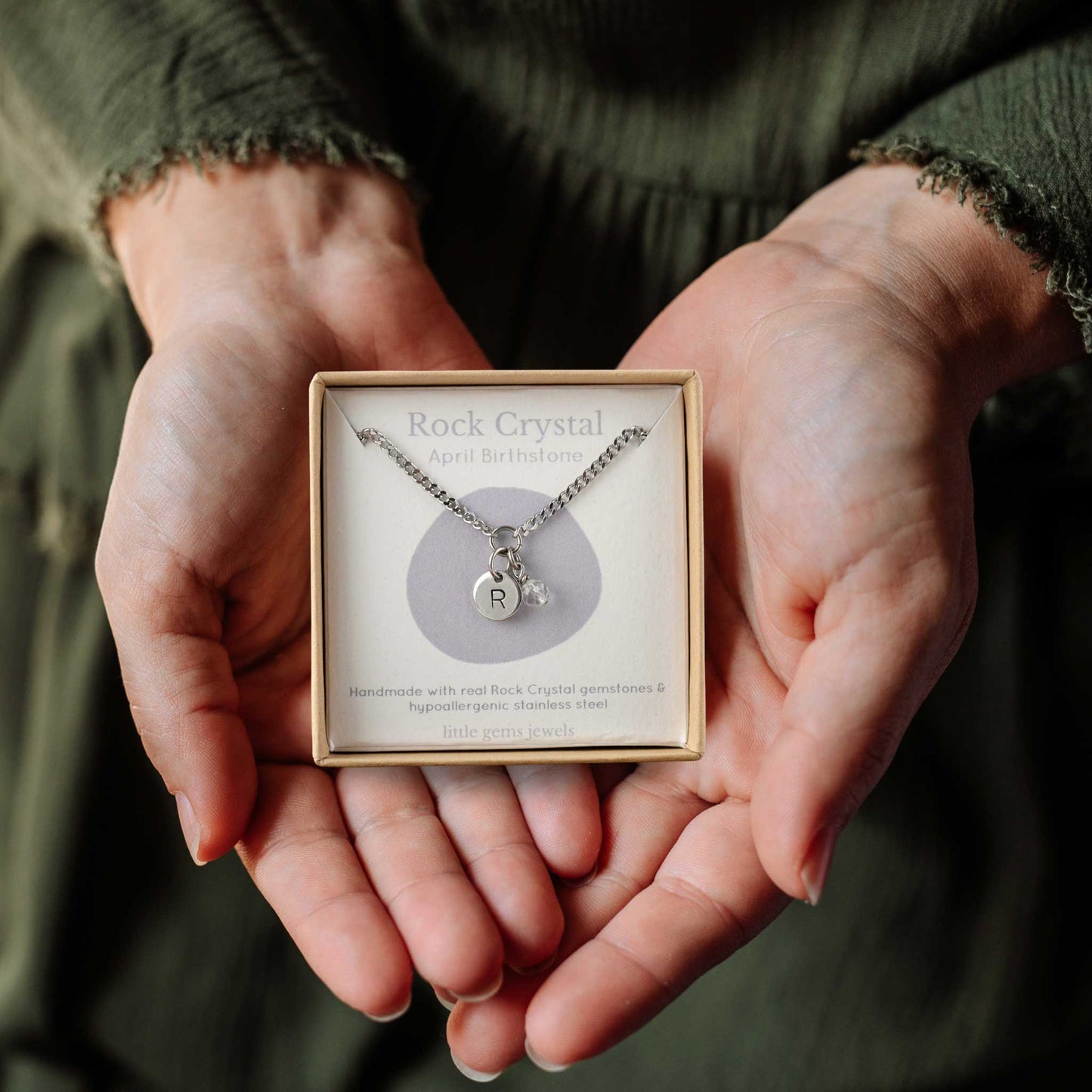 Hands holding a small square eco-friendly gift box containing a tiny initial and Rock Crystal April birthstone necklace