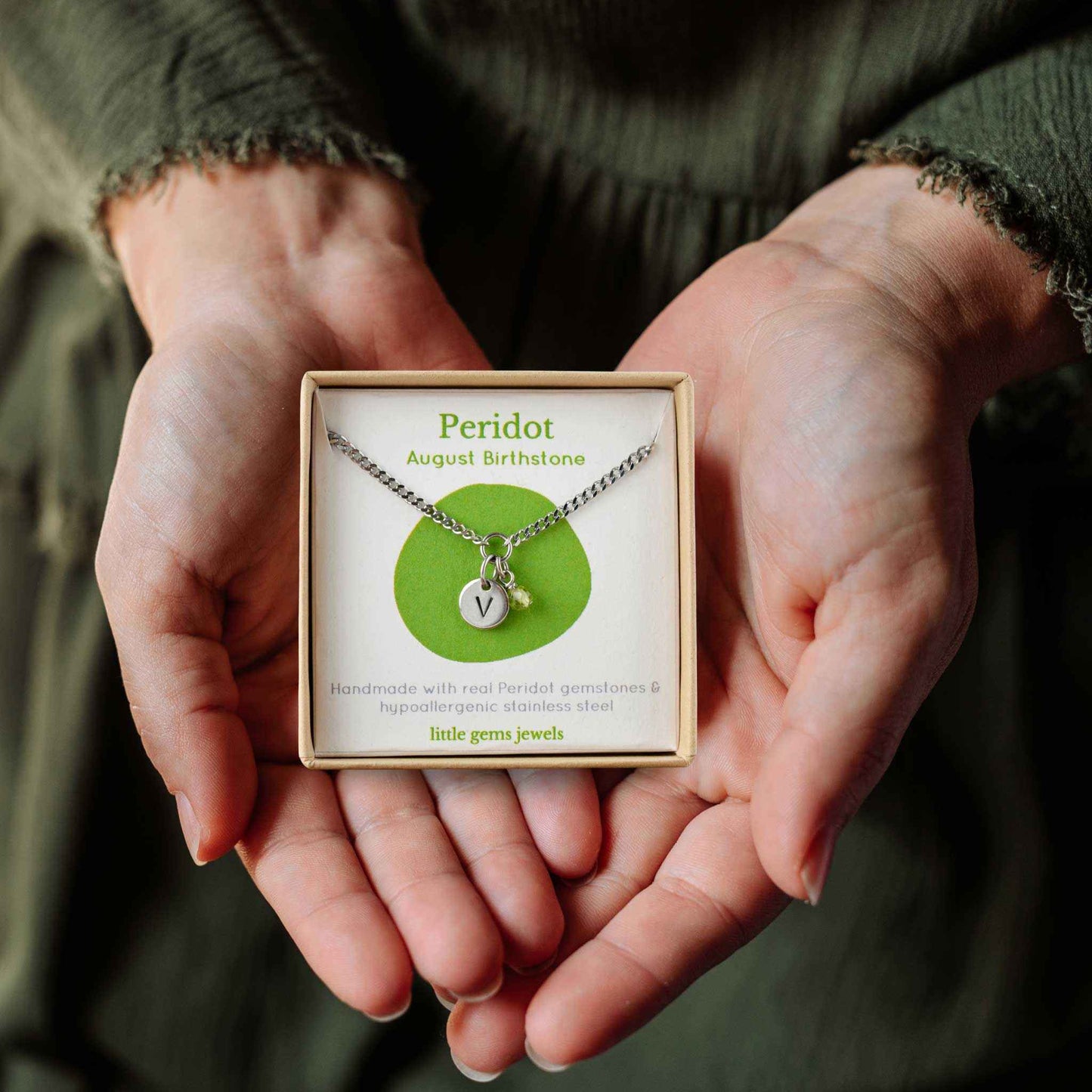 Hands holding a small square eco-friendly gift box containing a tiny initial and Peridot August birthstone necklace