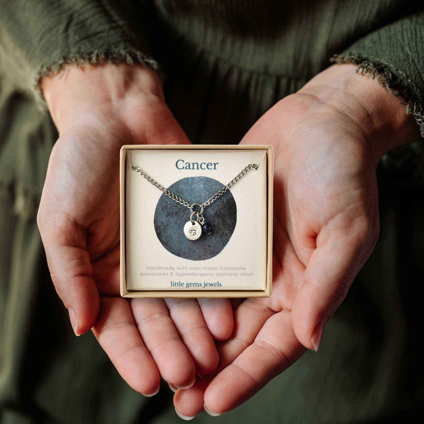 Hands holding a small square eco-friendly gift box containing a dainty Cancer zodiac sign necklace