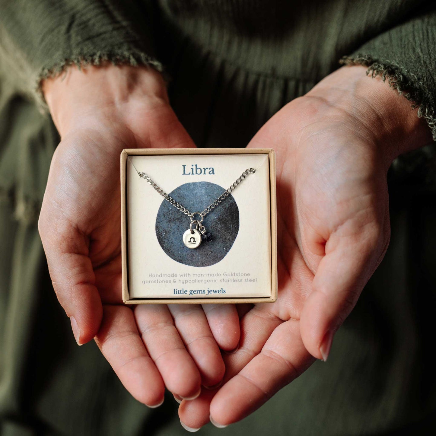 Hands holding a small square eco-friendly gift box containing a dainty Libra zodiac sign necklace