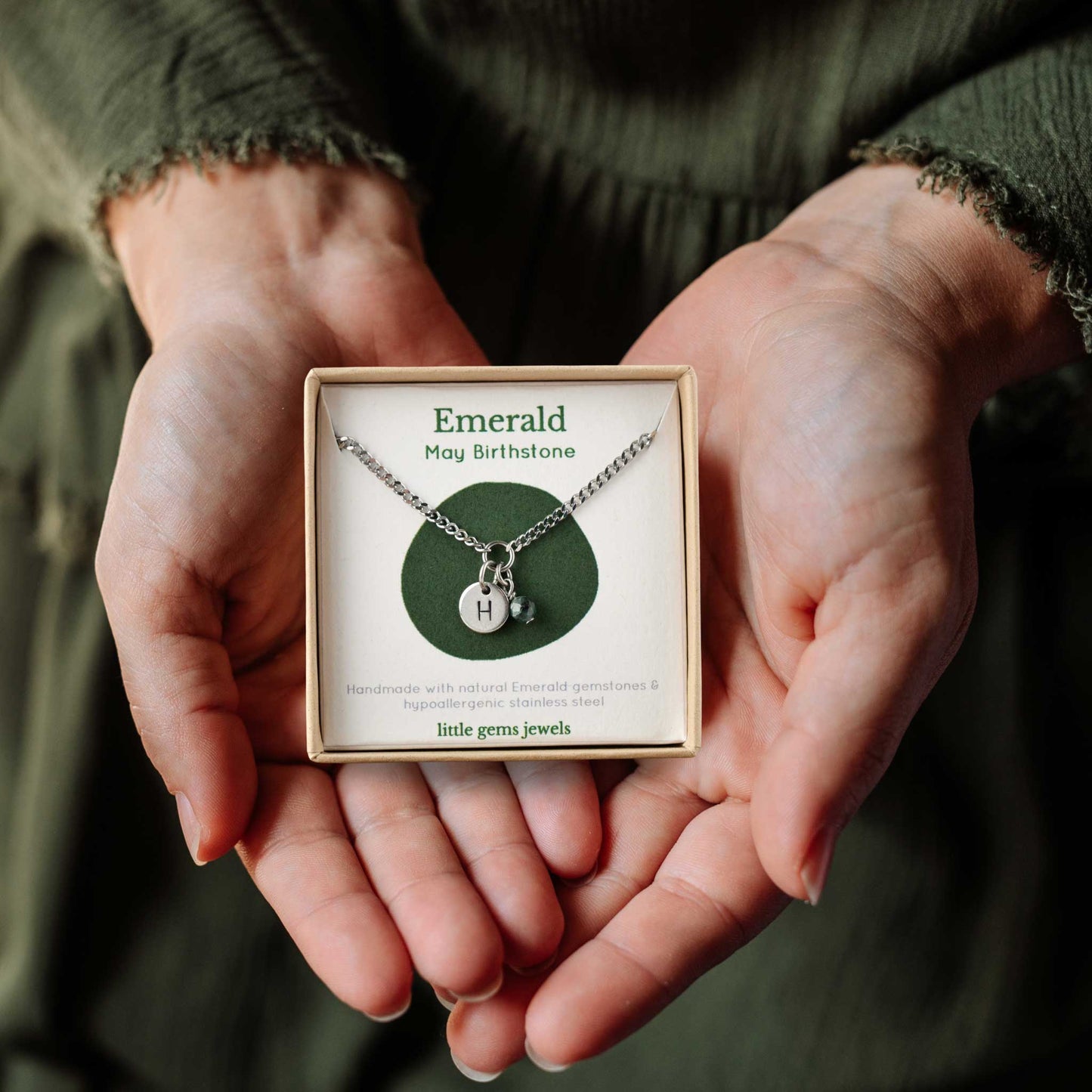 Hands holding a small square eco-friendly gift box containing a tiny initial and Emerald May birthstone necklace