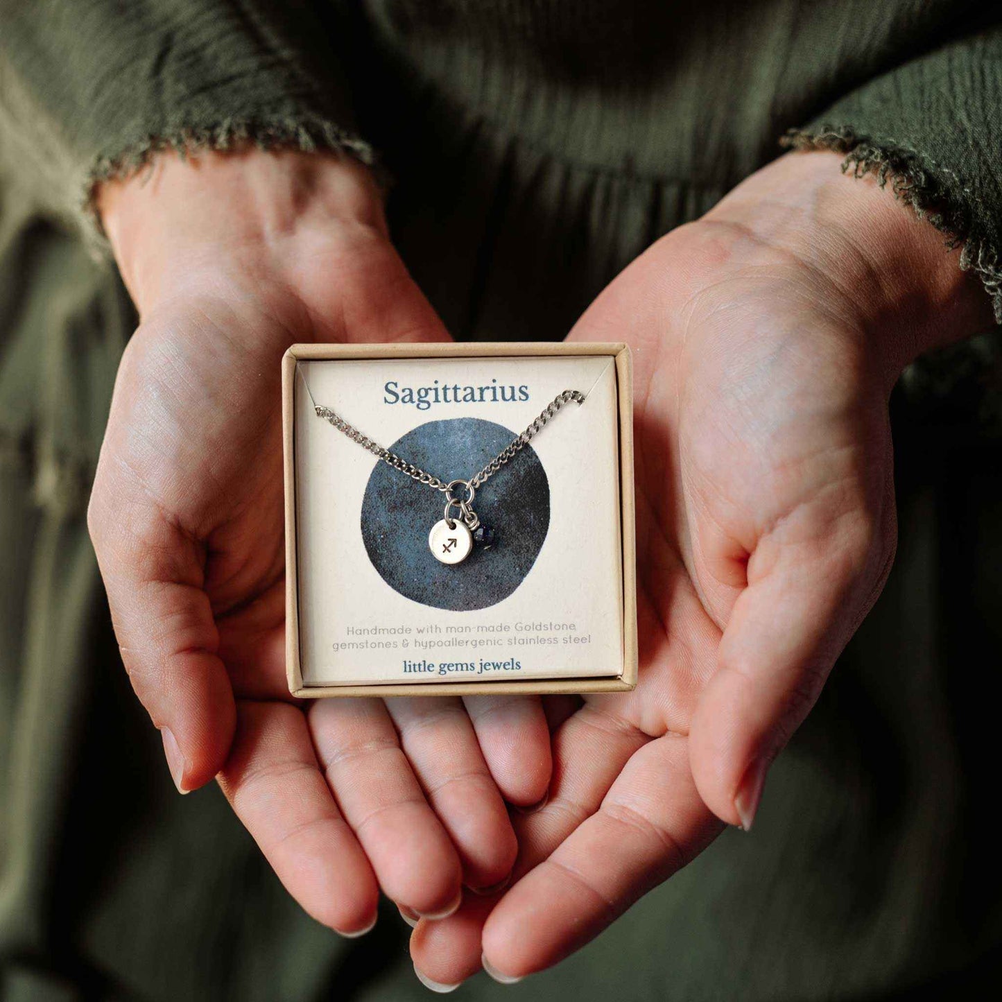 Hands holding a small square eco-friendly gift box containing a dainty Sagittarius zodiac sign necklace