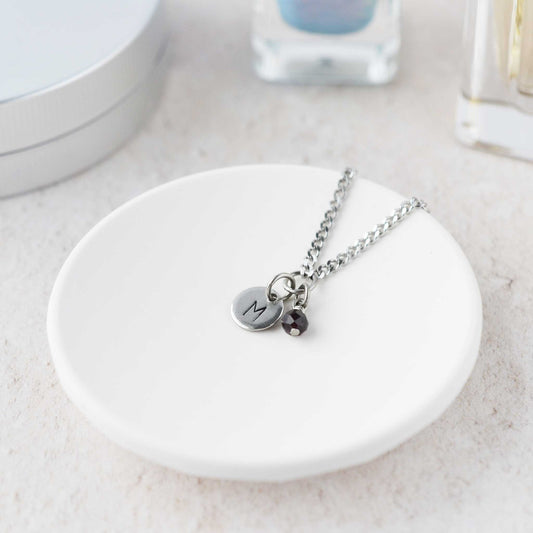 Tiny necklace with round disc pendant stamped with letter M and Garnet gemstone charm laying in a white trinket dish