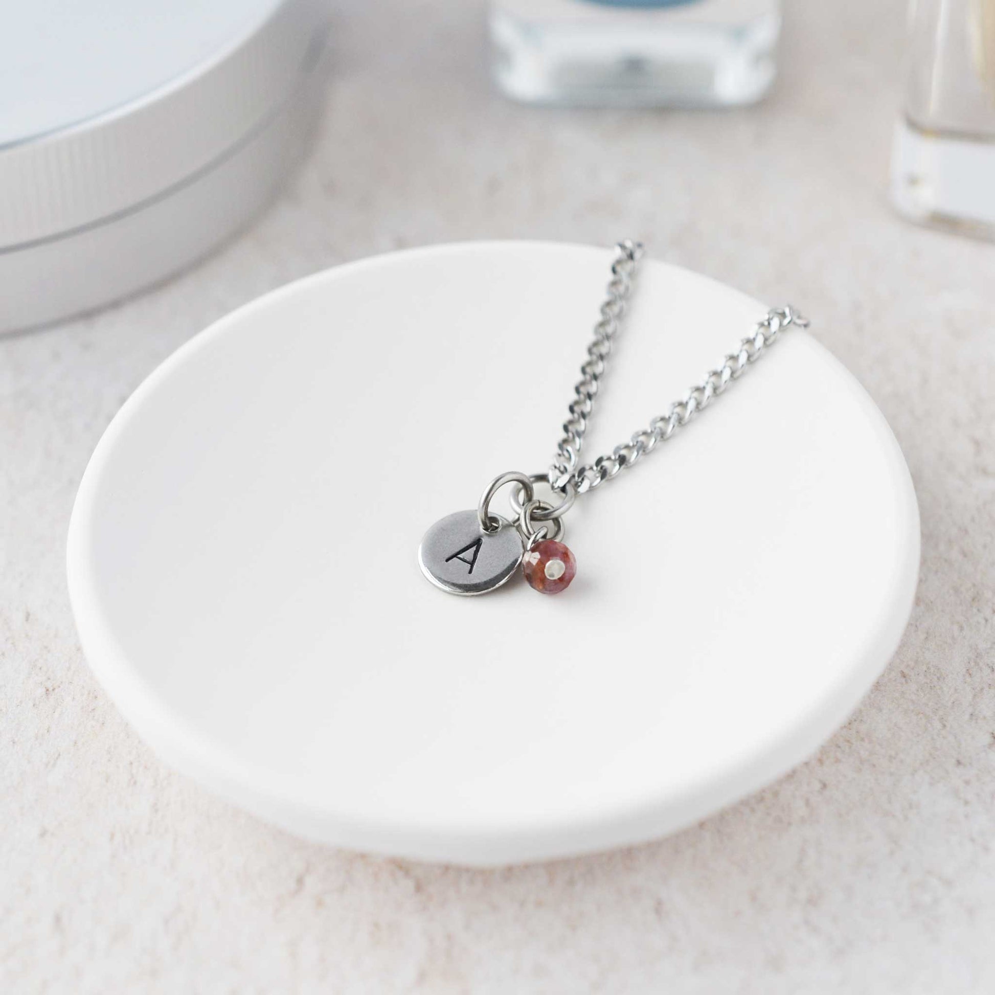 Tiny necklace with round disc pendant stamped with letter A and Ruby gemstone charm laying in a white trinket dish