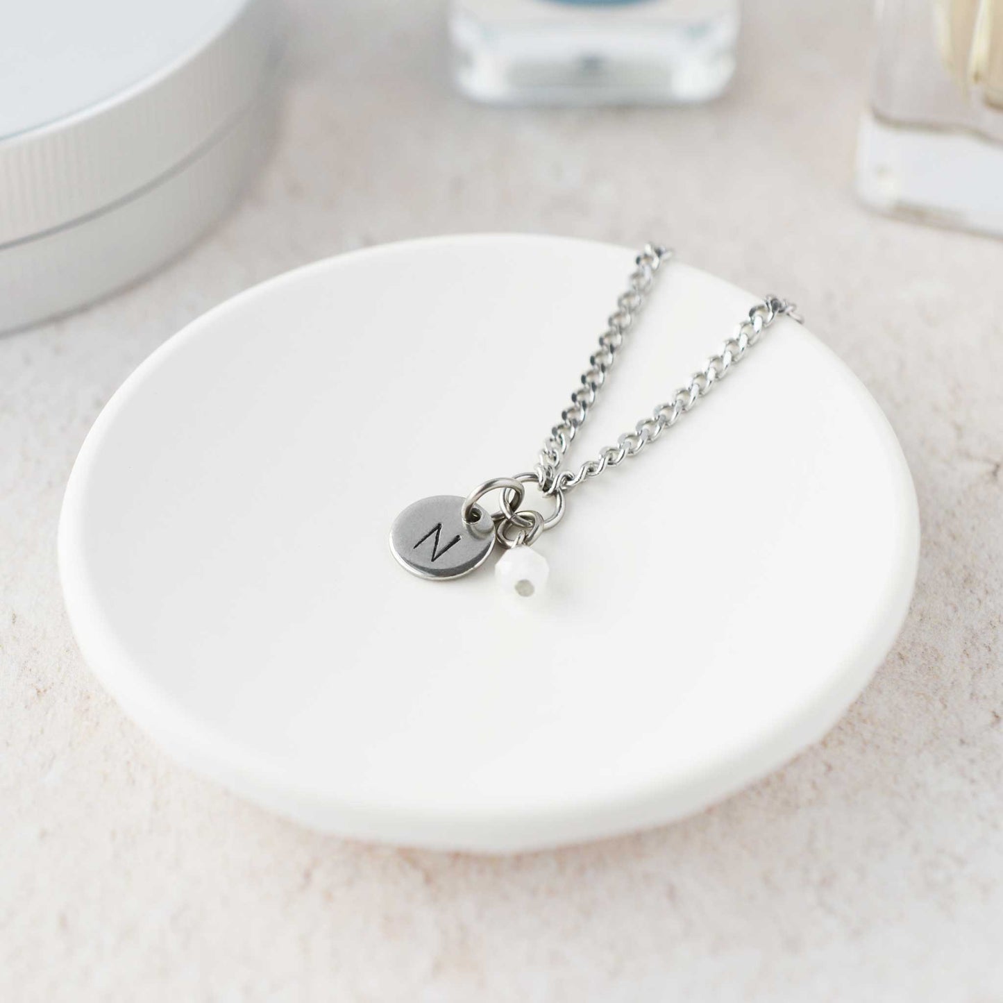 Tiny necklace with round disc pendant stamped with letter N and Moonstone gemstone charm laying in a white trinket dish