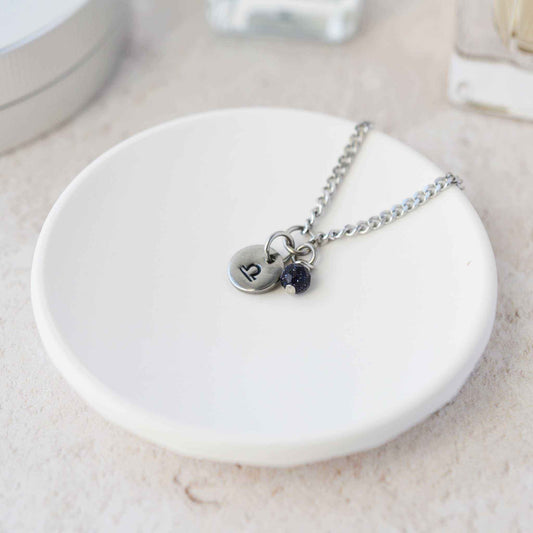 Tiny Libra zodiac sign disc pendant necklace with Blue Goldstone charm laying on white trinket dish