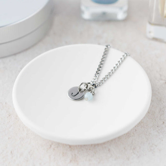Tiny necklace with round disc pendant stamped with letter J and Aquamarine gemstone charm laying in a white trinket dish