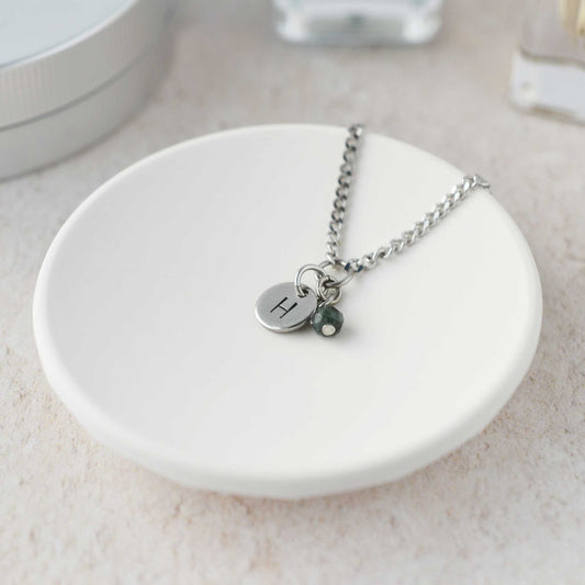 Tiny necklace with round disc pendant stamped with letter H and Emerald gemstone charm laying in a white trinket dish