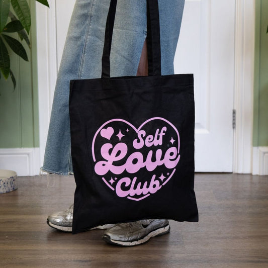 Black tote bag with 'Self Love Club' text held by a person wearing jeans and sneakers.