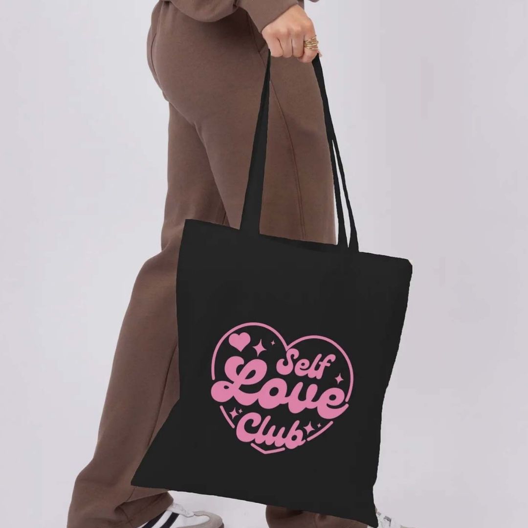 Black tote bag with pink 'Self Love Club' design held by a person in a brown outfit.