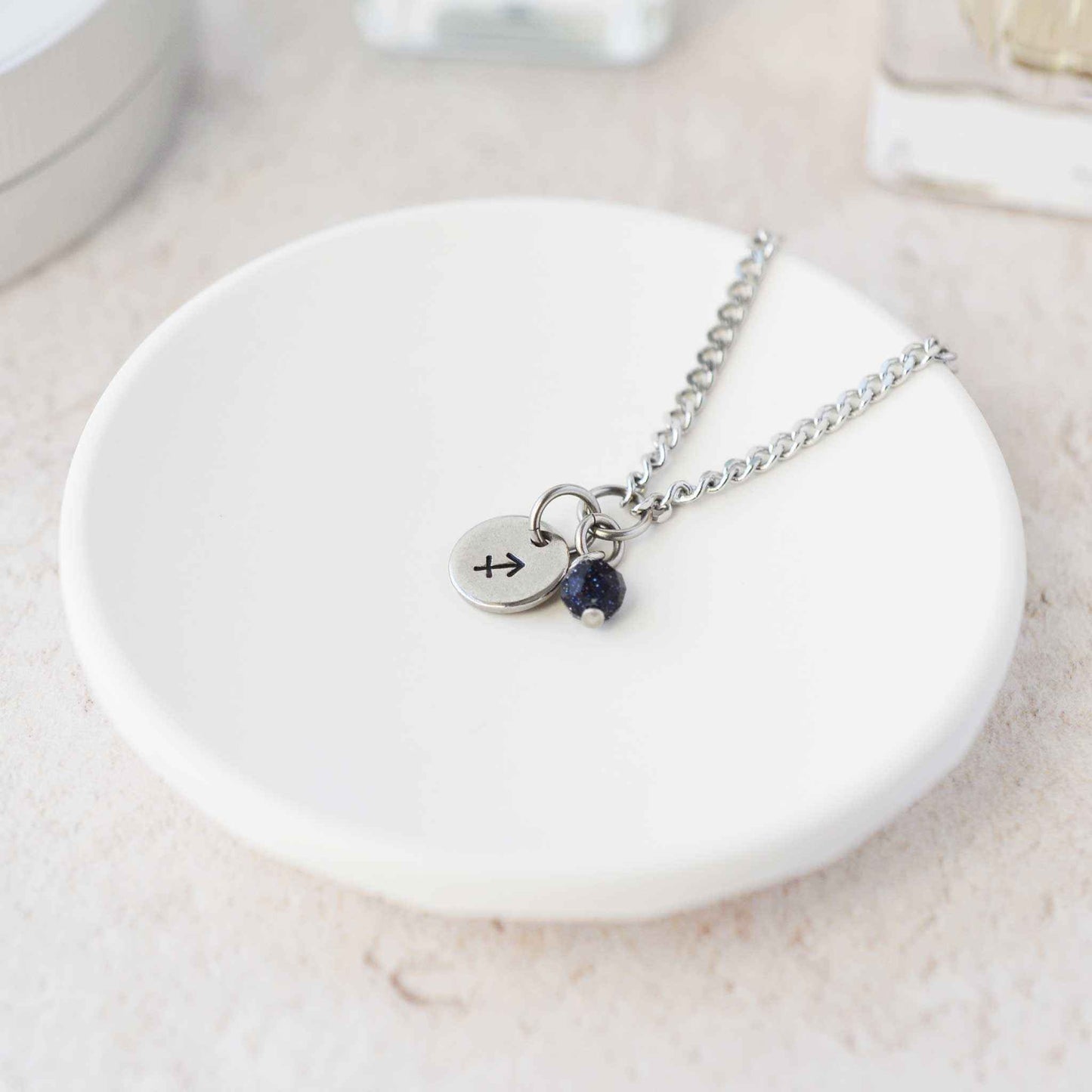 Tiny Sagittarius zodiac sign disc pendant necklace with Blue Goldstone charm laying on white trinket dish