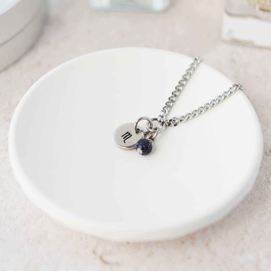 Tiny Scorpio zodiac sign disc pendant necklace with Blue Goldstone charm laying on white trinket dish