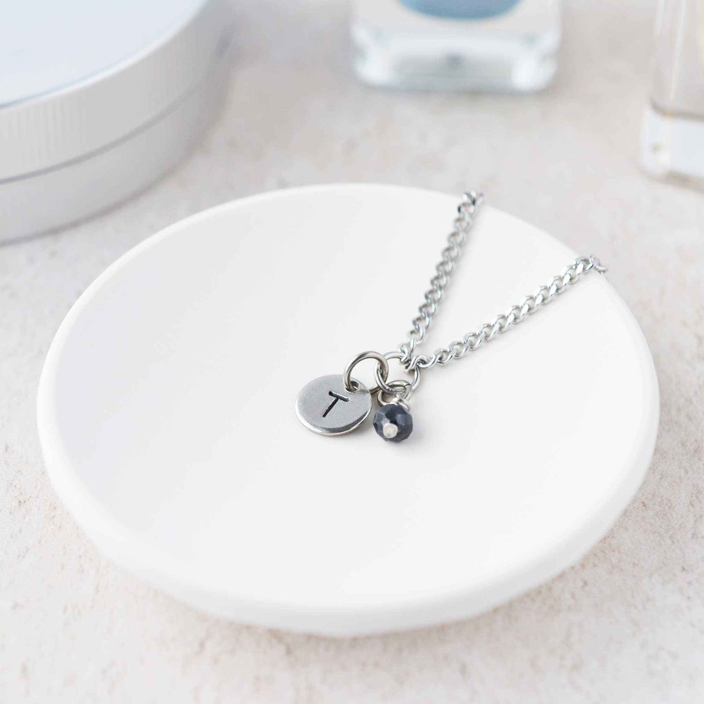 Tiny necklace with round disc pendant stamped with letter T and Sapphire gemstone charm laying in a white trinket dish