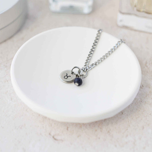 Tiny Taurus zodiac sign disc pendant necklace with Blue Goldstone charm laying on white trinket dish