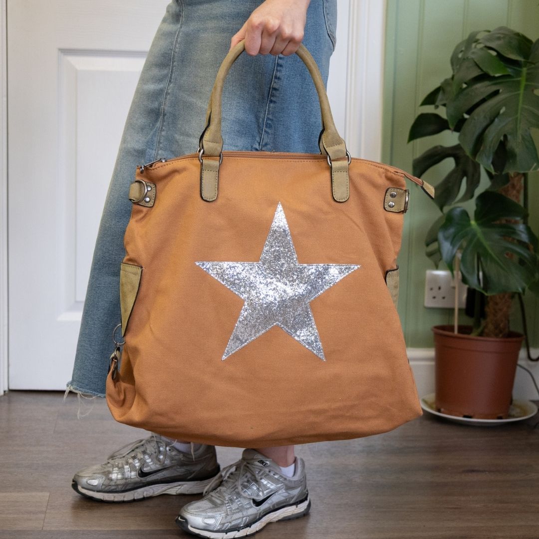 Person holding a brown tote bag with a silver star, wearing jeans and silver shoes, in a room with a plant.