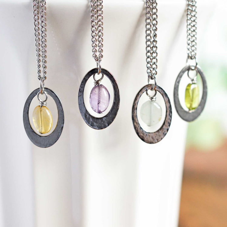 Four different coloured gemstone pendant necklaces hanging against white cup
