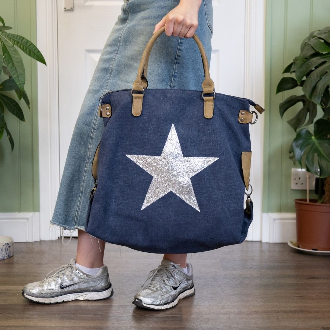 Woman wearing denim skirt and trainers holding navy tote bag with silver star emblem.