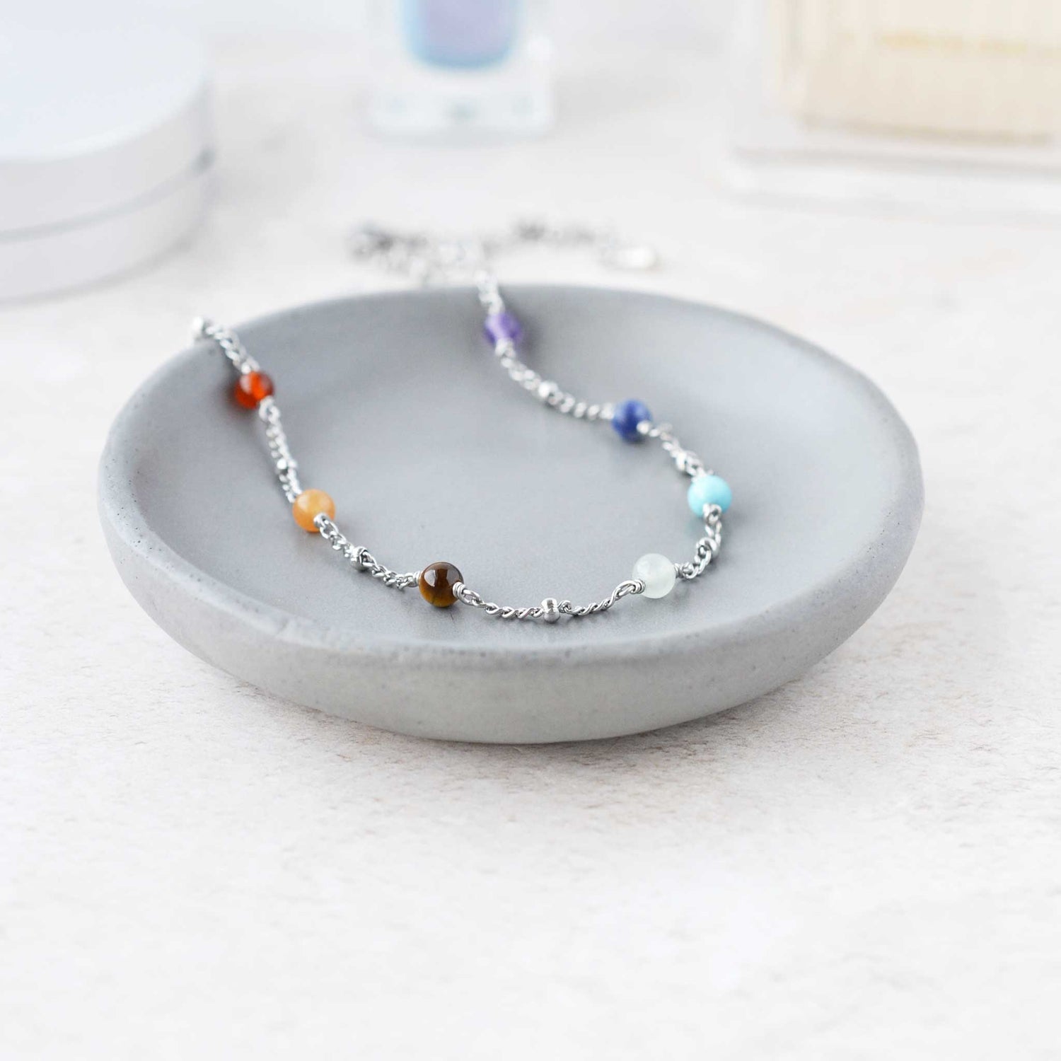 Gemstone chakra necklace lying on grey trinket dish on dressing table.