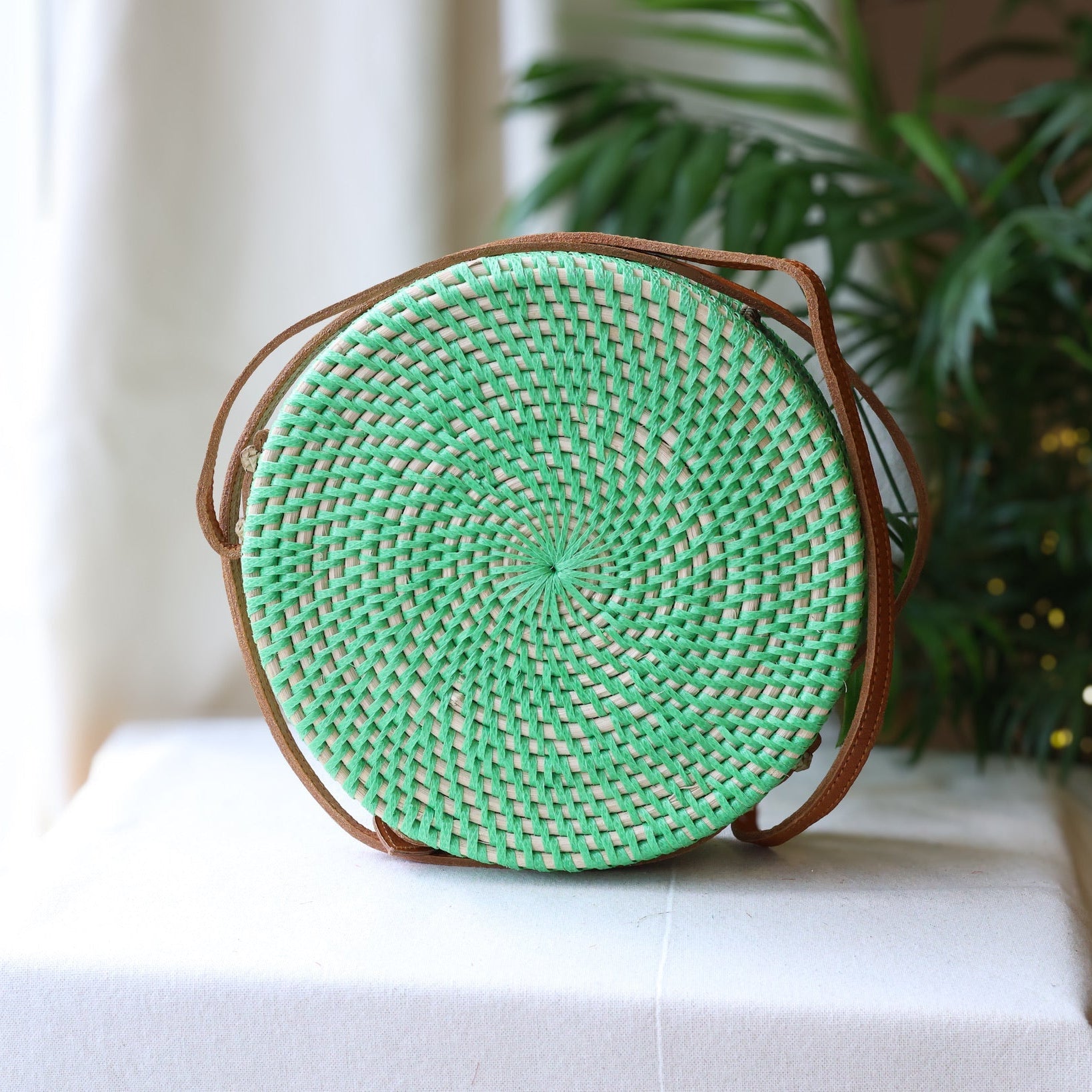 Green woven round bag on a white surface with a blurred plant in the background