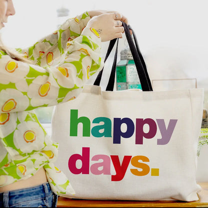 Person holding a tote bag with 'happy days' text in colorful letters.