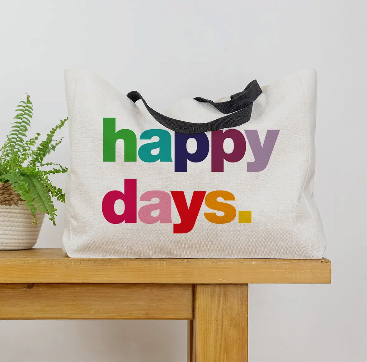 Tote bag with 'happy days' text in colorful letters on a wooden surface.