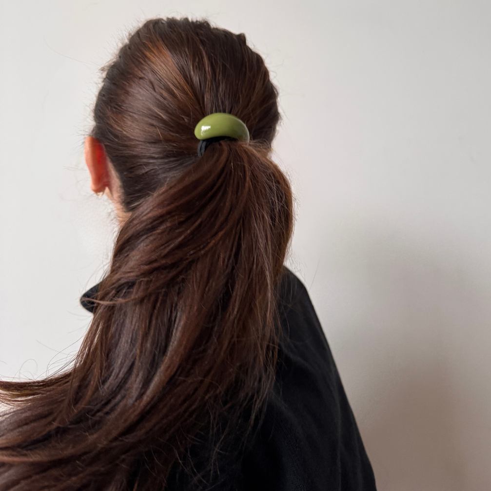 Person with brown hair tied in a ponytail using a green hair tie against a plain background