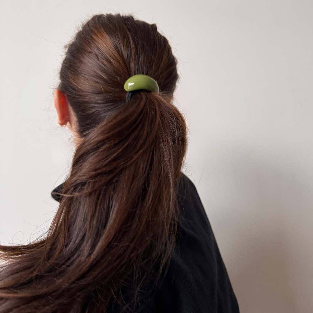 Person with brown hair tied in a ponytail using a green hair tie against a plain background