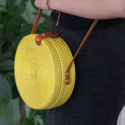Yellow woven handbag with brown strap held by a person against a green plant background