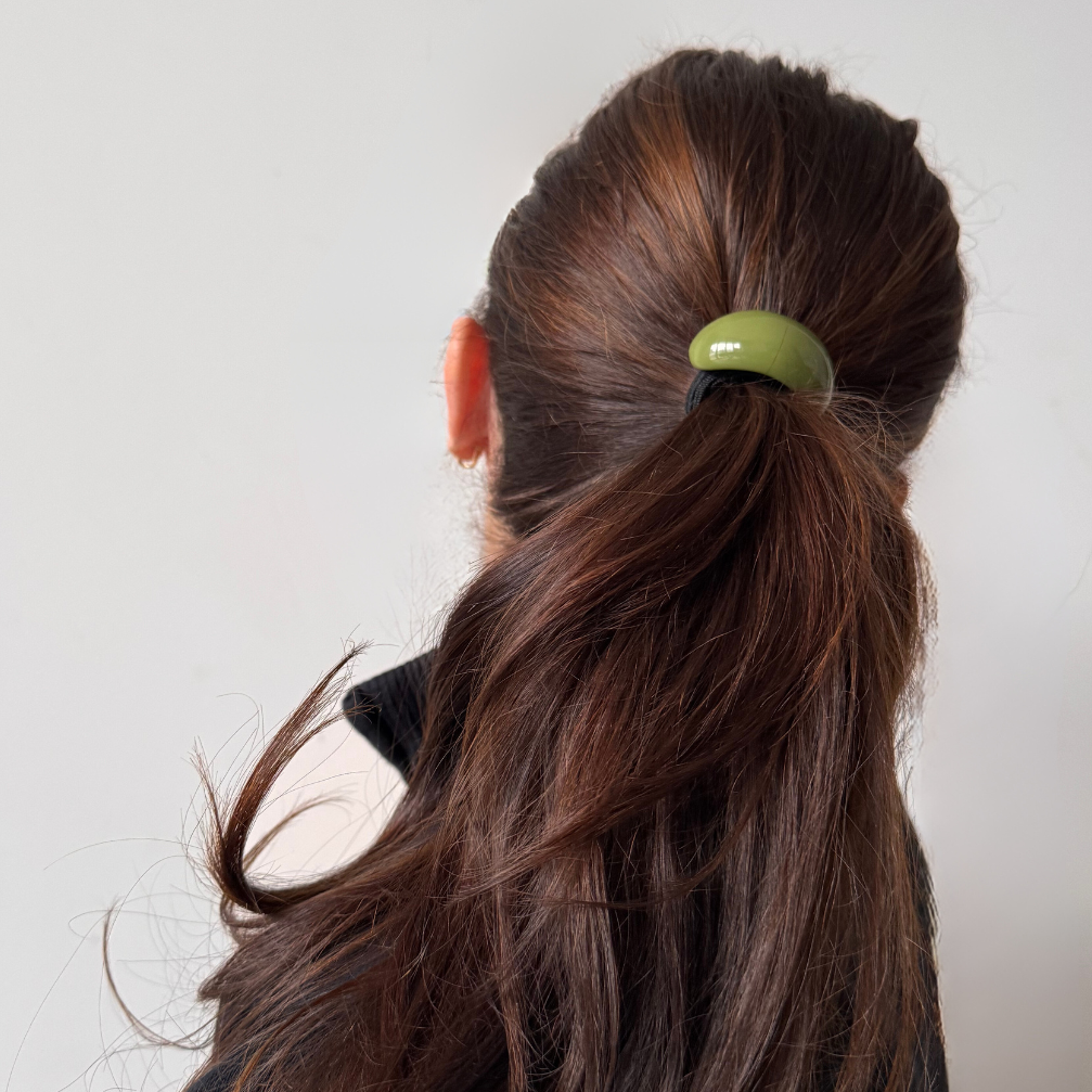 Person with long brown hair tied into a ponytail with a green hair tie against a plain background