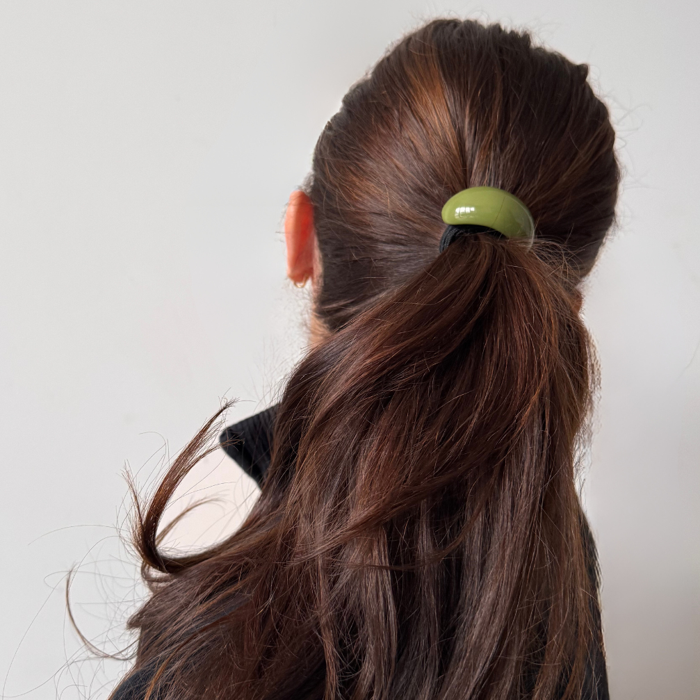 Person with long brown hair tied into a ponytail with a green hair tie against a plain background