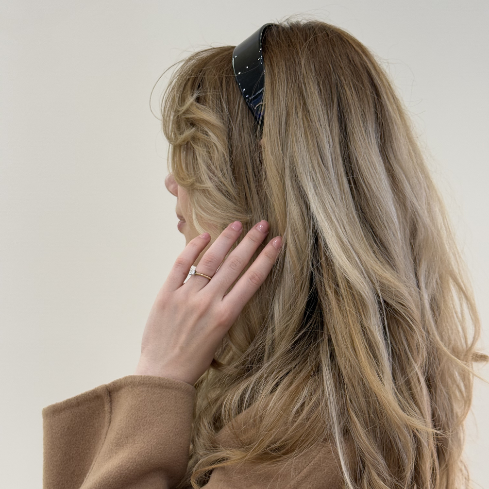 Person with long, wavy hair wearing a blck hairband and a brown coat against a plain background