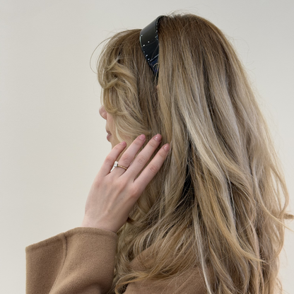 Person with long, wavy hair wearing a black hairband and a brown coat against a plain background
