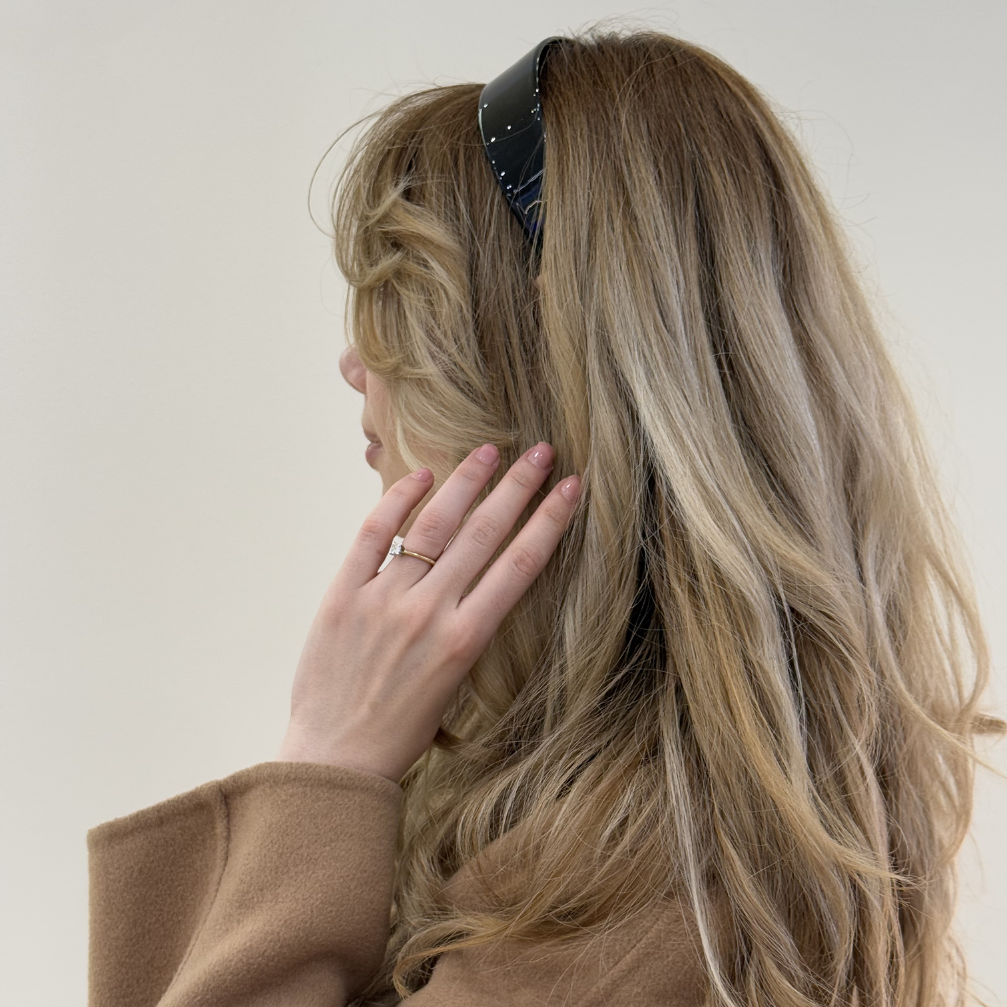 Person with long, wavy hair wearing a brown coat and a black headband against a plain background