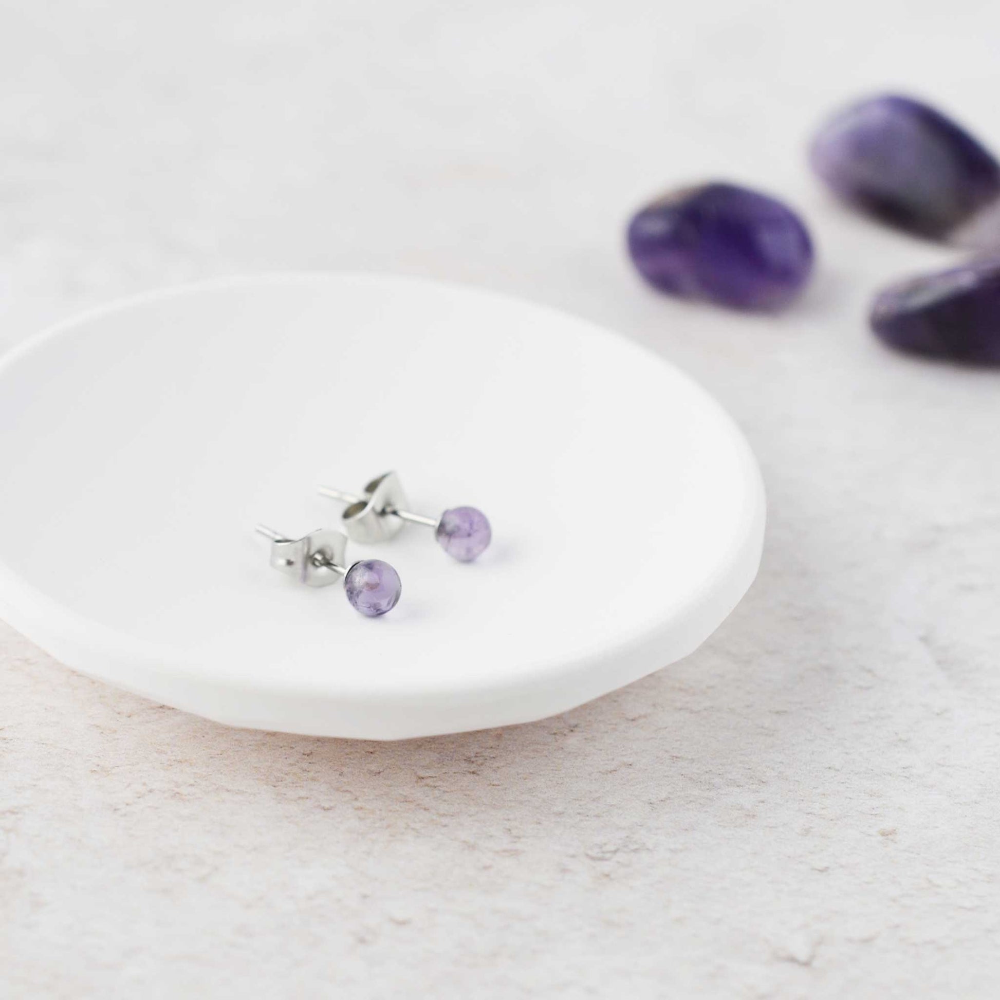 Tiny round purple Amethyst stud earrings in white trinket dish.